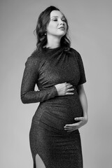 Black and white portrait of young pregnant female in grey sequin dress with hands near pregnant belly.