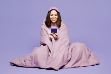 Wall Mural - Full body young calm woman wear pyjamas jam sleep eye mask rest relax at home sit wrapped in blanket hold use mobile cell phone isolated on plain pastel purple background. Good mood night nap concept.