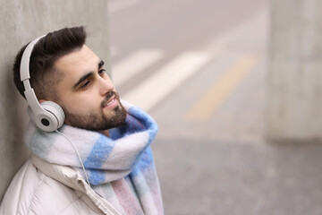 Wall Mural - Smiling man in warm scarf and headphones on city street. Space for text