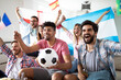 © NDABCREATIVITY - Diverse friends sports fans watching football match on TV at home. Celebrating shouting excited