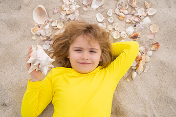 Kid laying on sandy beach with Shells. Summer kids. Portrait from above of a boy holding seashell laying on the sand. Summer holidays with children.