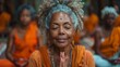 © familymedia - An older woman with grey hair is meditating with her hands in a prayer position surrounded by other participants in orange attire