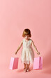 © LIGHTFIELD STUDIOS - A little girl in a white dress happily holds shopping bags against a pink backdrop.