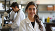 © easybanana - A young scientist wearing a lab coat smiles at the camera while her colleague works in the background.