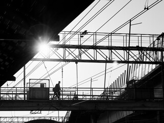 A lone person walking on an urban bridge silhouetted by the setting sun, surrounded by utility lines and structures.