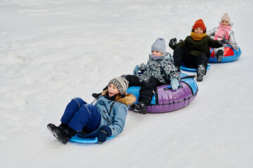 Row of adorable happy children in winter jackets sitting on slides and snow tubes and riding down hill in natural environment