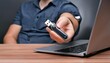 © blackdiamond67 - A man holds a flash drive in his hand against the background of a laptop lying on a wooden table.
