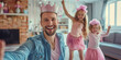 © MNStudio - Young cheerful father wearing princess tiara while playing with his two small daughters. Parent and two little girls wearing pink clothes at home.