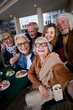 © CarlosBarquero - Vertical. Happy mature gray hair woman taking selfie of group seniors Caucasian cheerful friends posing together sitting at cafeteria. Older people looking laughing at camera with snacks on table