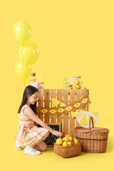 Canvas Print - Cute little Asian girl with lemons near lemonade stand on yellow background