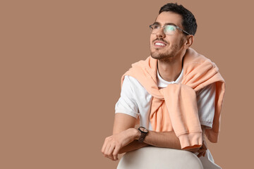Wall Mural - Handsome young man in stylish clothes with eyeglasses sitting on chair and posing against beige background