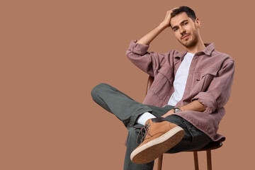 Wall Mural - Handsome young man in stylish clothes sitting on stool and posing against beige background