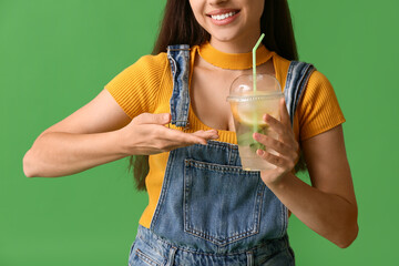 Canvas Print - Beautiful young happy woman pointing at cup of fresh lemonade on green background, closeup