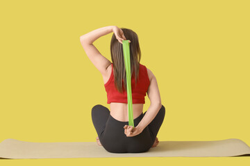 Wall Mural - Sporty young woman with stretching band doing yoga on mat against yellow background, back view