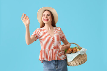 Poster - Beautiful young happy woman with food for picnic in wicker basket waving hand on blue background