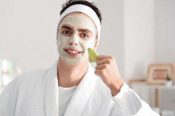 Poster - Young man with applied clay mask and facial massage tool in bathroom, closeup