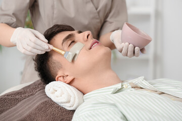 Poster - Beautician applying clay mask on young man's face in salon, closeup