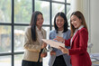 © amnaj - Three businesswomen discuss work while holding documents, standing in a modern office with large windows, smiling and engaged.