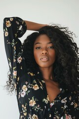 Wall Mural - young Black woman standing with her arm raised, wearing a black floral dress, her long curly hair flowing, natural light casting shadows, white background