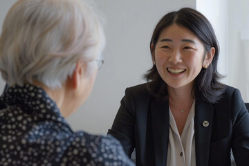 Wall Mural - Japanese female professional in business attire, smiling and speaking to an elderly woman during the interview process at her office desk. The background is white with soft lighting, creating a comfor