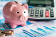 © lashkhidzetim - A pink piggy bank sits on top of a calculator and a stack of dollar bills. Concept of saving money