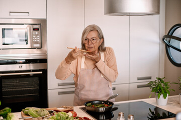 Smiling woman at home tasting food from the pan with a table spoon. People in the kitchen preparing dinner. One elderly female alone cooking. Real lifestyle person testing taste and cook preparation.