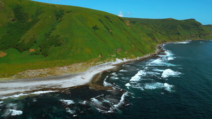 Wall Mural - Wild nature, green mountains with steep green slopes leading to the sea. Clip. Aerial of foam of blue waves break in volcanic rocks.