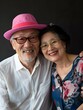© Pascal - Elegant senior Asian couple with smiles, man wearing a pink hat and glasses, woman in chic blouse, embodying fashionable aging and joyful companionship