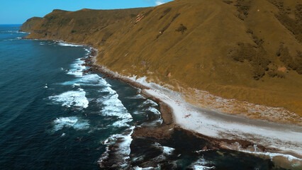 Wall Mural - Aerial of mountain covered by burnt yellow grass and blue sea. Clip. Steep slope of yellow hill and dark blue waves crashing in stones and creating foam.