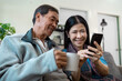 © itchaznong - Elderly married couple who are retired are relaxing together in living room, using mobile