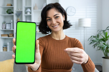 Close-up portrait of a young woman sitting at home, holding and showing a mobile phone to the camera, pointing at the green screen