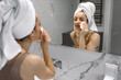 © Nataliya - Charming young woman with white towel on her head standing in the bathroom and wiping the skin on her face while looking in the mirror. Care, health, lifestyle concept