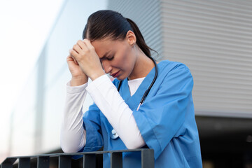 Tired exhausted young female nurse or doctor standing outside hospital, crying and feeling stressed after hard working day