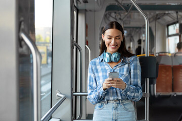 Happy European woman with headphones using cellphone, training in modern tram, enjoying comfortable public transport. Portrait of passenger with phone and headset