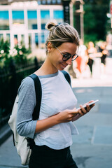 Wall Mural - smiling hipster girl using smartphone for searching cafes in downtown strolling on street,positive f