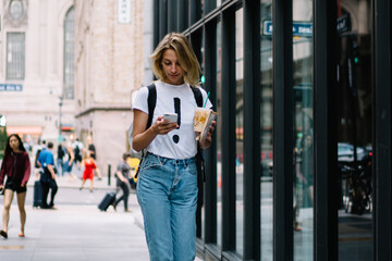 Wall Mural - Young female tourist walking on city street with coffee takeaway checking route on mobile application, casually dressed hipster girl reading income message on smartphone strolling in downtown