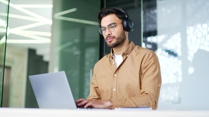 Confident IT specialist developer in wireless headphones works on laptop computer sitting at workplace at desk in business office. Serious focused coder in glasses is writing code, works on a project
