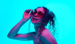© master1305 - Portrait of beautiful young woman with bare shoulder, curly hair in ponytail, posing in sunglasses against cyan background in neon light. Concept of beauty, wellness, self-care, fashion, cosmetics