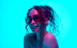 © master1305 - Portrait of beautiful young woman with bare shoulder, curly hair in ponytail, posing in sunglasses against cyan background in neon light. Concept of beauty, wellness, self-care, fashion, cosmetics