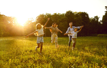 Happy family running and having fun in summer park enjoying sunny day in nature. Mother, father and their cheerful children boy and girl walking on green grass outdoors at sunset together.