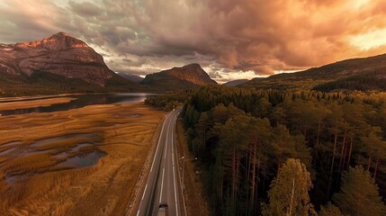 Canvas Print -   An aerial shot of a road winding through a dense forest, surrounded by towering mountains and reflecting on a serene body of water in the foreground