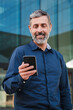 © Jose Calsina - Vertical portrait. Real mature male worker watching a finance app on his cellphone. Middle aged person holding a mobile phone smiling. Handsome mid adult man smiling using a smartphone at workplace.