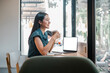 © Satori Studio - A woman is sitting at a desk with a laptop and a cup of coffee. She is smiling and she is enjoying her coffee and work