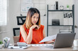 © Satori Studio - A woman in an orange shirt is sitting at a desk with a laptop and a stack of papers. She is deep in thought, possibly working on a project or analyzing data. Concept of focus and concentration