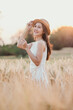 © Satori Studio - A woman is standing in a field of tall grass, wearing a white dress and a straw hat. She is smiling and holding a bunch of flowers in her hand. Concept of peace and relaxation