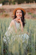 © Satori Studio - A woman is sitting in a field of tall grass, wearing a straw hat and a white dress. She is smiling and looking at the camera. Concept of relaxation and enjoyment of nature