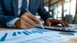 © admin_design - Business professional analyzing financial graphs on a paper while working on a laptop in a modern office environment.