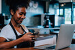 © sorapop - Close up portrait of focused black woman coffee shop owner in apron using smartphone in her cafe. Multi tasking barista manages orders communicates with customers runs her own business.