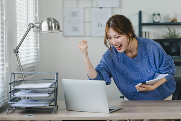 Triumphant Asian businesswoman celebrating professional victory with arms raised in front of a laptop at home office.