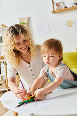 Wall Mural - Curly-haired mother gently guides her toddler daughters hand while drawing with colorful crayons at home.